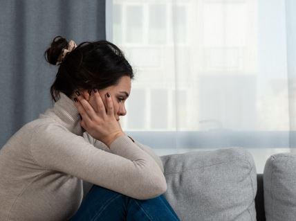 Devojka u bež rolci se drži rukama za glavu na sivom kauču i gleda dole