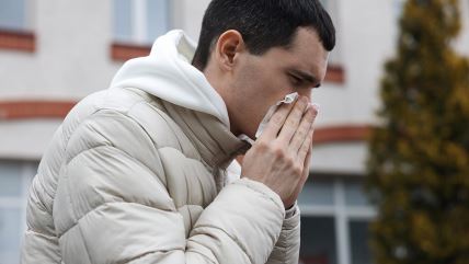 Muškarac u bež jakni napolju duva nos u maramicu