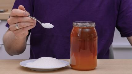 Ruka drži kašičicu šećera iznad tanjira sa šećerom i pored tegle meda