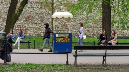 Prodavac u Krakovu gura plava kolica sa uličnom hranom puna pereca, dok drugi sede na klupama, razgovaraju ili koriste telefone